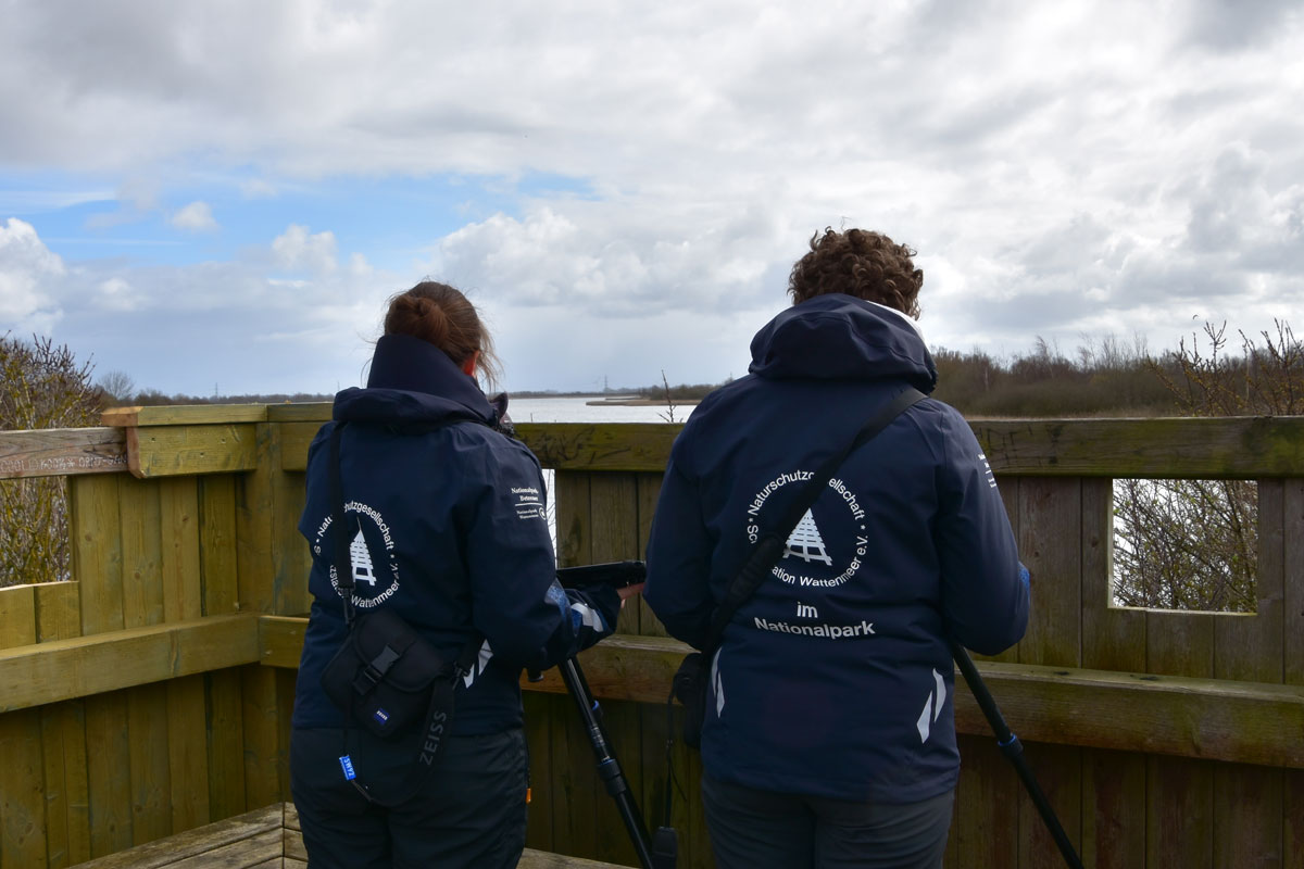 zwei Menschen mit Jacken der Schutzstation Wattenmeer auf einer Aussichtsplattform.