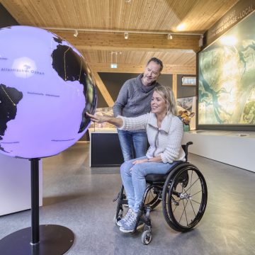 Eine Frau im Rollstuhl zeigt auf einen Globus, ein Mann steht neben ihr.