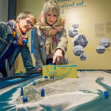 Zwei Frauen betrachten das Model der Insel Wangerooge. Sie bedienen einen Hebel um den einen Querschnitt aus dem Modell empor zu heben.